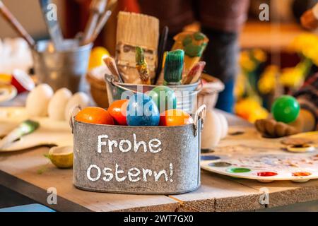 16. März 2024: Happy Easter-Schriftzug auf einem Metallkorb mit bunten Eiern auf einem rustikalen Tisch mit Eierfarbe. FOTOMONTAGE *** Frohe Ostern Schriftzug auf einem Korb aus Metall mit bunten Eiern auf einem rustikalen Tisch mit Eierfarbe. FOTOMONTAGE Stockfoto