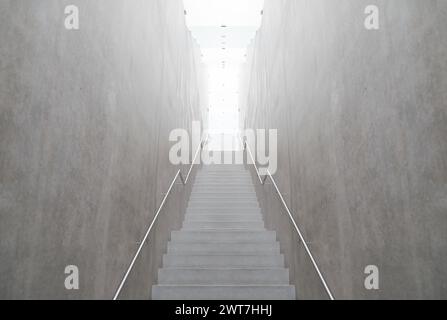 Leere, schmale Treppe, die zum Licht führt. Betonwände und beleuchtete Decke aus satiniertem Glas. Handläufe auf beiden Seiten. Stockfoto