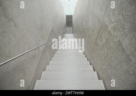 Leere schmale Treppe nach unten. Betonwände und beleuchtete Decke aus satiniertem Glas. Handlauf auf der linken Seite. Foto in Grautönen. Stockfoto