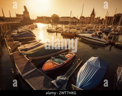 Sonnenuntergang im Hafen von Lindau. Orangefarbenes Bild von zahlreichen verankerten Booten und Segelbooten. Niedrige Sonne und lange Schatten, perspektivische Sicht auf den gekrümmten Steg. Stockfoto