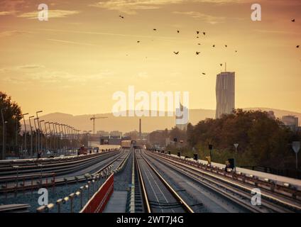 Erhöhte U-Bahn-Linie in Richtung Donaucity - schwindende Perspektive. Sonnenuntergang in der Stadt, Berge am Horizont. Vogelherde, die überfliegen. Stockfoto