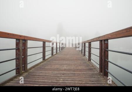 Eine hölzerne Promenade mit Handgeländern über einem See im Nebel. Holzsteg über stilles Wasser. Der Laufweg verläuft vom Betrachter nach links. Stockfoto