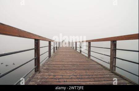 Eine hölzerne Promenade mit Handgeländern über einem See im Nebel. Holzsteg über stilles Wasser. Der Gehweg verläuft vom Betrachter nach rechts. Stockfoto