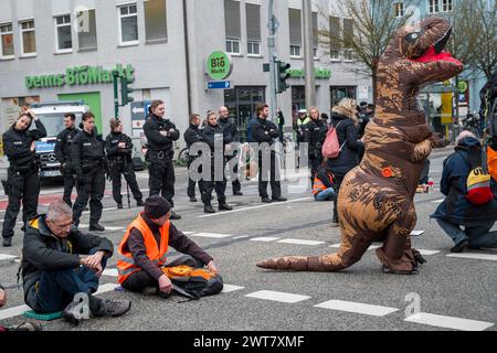 Regensburg, Deutschland. März 2024. Demonstranten, die in der Straße sitzen, blockieren die Kreuzung Kumpfmühler Str. und Fritz-Fend-Straße. Eine Person in einem Dinosaurierkostüm läuft die Straße entlang. Polizeibeamte stehen in der Nähe. Die letzte Generation protestiert gegen die Klimapolitik der Ampelregierung. Quelle: Daniel Vogl/dpa/Alamy Live News Stockfoto