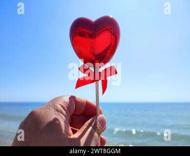 Ein Stock in Form eines volumetrischen roten Herzens in der Frauenhand vor dem Hintergrund des blauen Meeres, des Sandstrandes und des blauen Himmels an sonnigen Sommertagen. Concept Love, Valentinstag, Verlieben. Vertikal Stockfoto