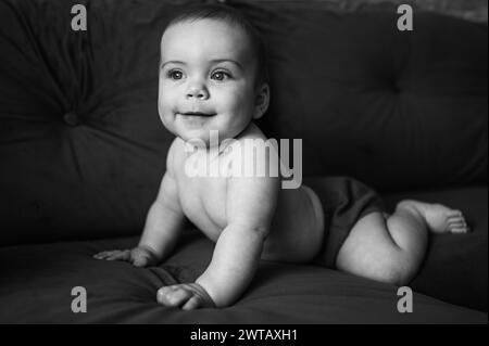 Niedliches emotionales lächelndes, lustiges Kind, das auf geraden Armen auf dunkelgrauem dunkelblauem Sofa sitzt. Baby-Gesichtsausdruck. Gesundes Kind. Haus komfortable Möbel, fröhliches Familienkonzept. Stockfoto