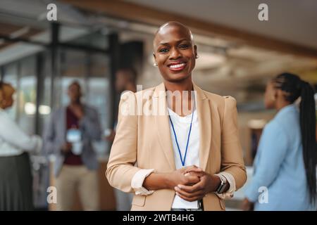 Strahlende professionelle afroamerikanische Frau lächelt selbstbewusst in einem Geschäftsumfeld, während Kollegen im Soft-foc Gespräche führen Stockfoto