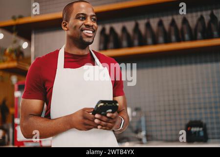 Fröhlicher Unternehmer, der in einem Café eine Pos-Maschine hält und eine weiße Schürze trägt. Ein Mann lächelt, während er sein Geschäft digital verwaltet, und repräsentiert kleine Bu Stockfoto