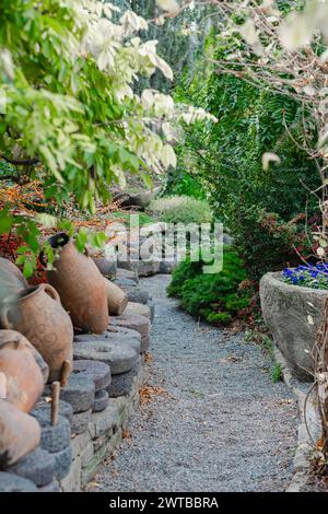 Ein ruhiger Kiesweg schlängelt sich durch einen üppigen Garten, flankiert von antiken Töpferwaren und lebendigen Pflanzen. Es lädt zu einem friedlichen Spaziergang im Grünen ein. Stockfoto