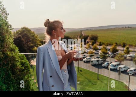 Eine Frau mittleren Alters steht in einem Straßencafé mit Blick auf die Berge bei Sonnenuntergang. Sie trägt eine blaue Jacke und trinkt Kaffee, während sie bewundert Stockfoto