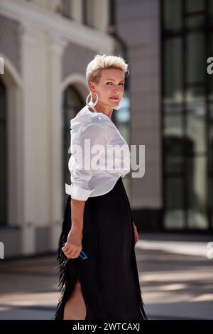 Stilvolle Fashionista mittleren Alters, gekleidet mit trendigem schwarzen Rock und einer weißen Bluse, schlendert durch die Straße und blickt mit Eleganz zurück. Schöne Reife Stockfoto