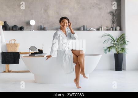 Lächelnde schwarze Frau im Bademantel, die am Rand einer luxuriösen freistehenden Badewanne sitzt Stockfoto