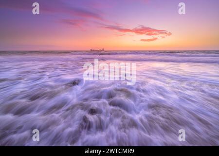 Wellen krachen am Strand in den Niederlanden während eines wunderschönen Sonnenuntergangs mit rosa und lila Farben an einem ruhigen Sommertag Stockfoto