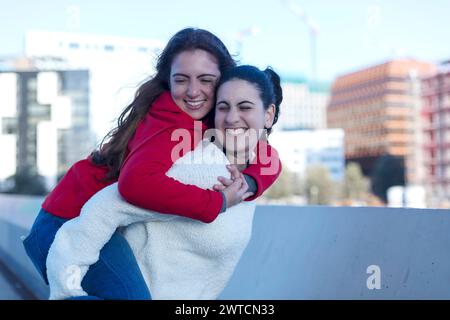 Zwei Freunde teilen sich einen fröhlichen Huckepack-Moment, einer in einem roten Hoodie, der andere in einer weißen flauschigen Jacke, beide lächeln und haben Spaß Stockfoto