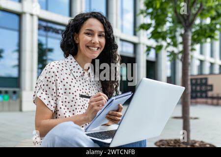 Eine unabhängige junge Frau in lässiger Geschäftskleidung konzentriert sich auf ihre Arbeit auf einem Laptop, während sie an einem hellen, sonnigen Tag draußen sitzt. Stockfoto
