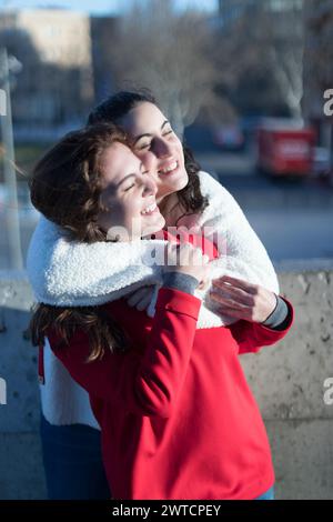 Zwei junge Frauen teilen sich eine fröhliche Umarmung im Freien, eine in einer weißen flauschigen Jacke und die andere in Rot, beide lächeln hell im Sonnenlicht Stockfoto