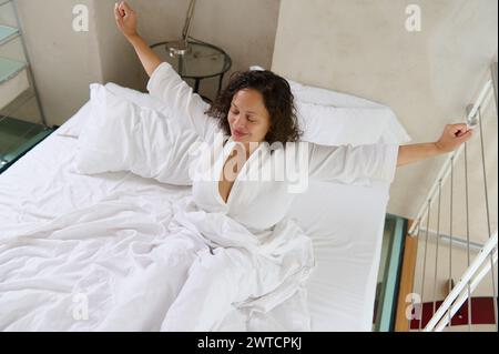 Blick von oben auf eine glückliche junge Frau, die nach dem Schlafen aufwacht und sich auf dem Bett ausdehnt. Blick aus einem hohen Winkel auf die wunderschöne Dame, die auf dem Bett lag und sich nach dem Aufwachen ausdehnte Stockfoto