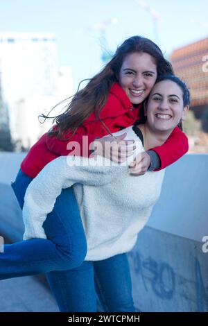 Zwei Freunde teilen sich einen fröhlichen Huckepack-Moment, einer in einem roten Hoodie, der andere in einer weißen flauschigen Jacke, beide lächeln und haben Spaß Stockfoto