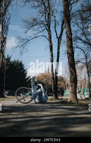 Ein aktiver Tag nimmt eine unerwartete Wende, während ein Radfahrer auf einen Parkweg stürzt, mit umgekipptem Fahrrad, inmitten der Ruhe von blattlosen Bäumen. Stockfoto