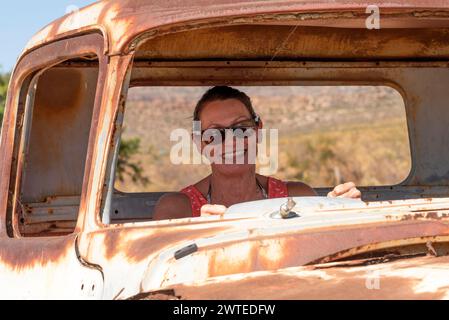 Südafrika. 16/02/2024. Attraktive Frau posiert mit einem alten rostigen amerikanischen Auto am Straßenrand in der Swartland-Region Südafrikas. Stockfoto