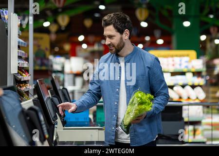 Erwachsener Mann in einem blauen Hemd mit einem Selbstkassenautomaten in einem Supermarkt, der frischen Salat hält. Stockfoto