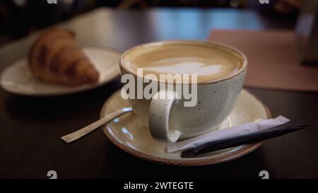 Köstlicher heißer italienischer Cappuccino steht in einer Tasse mit einem Zuckerpas auf einem Holztisch im Café. Im Hintergrund ein frisches Croissant auf einem Teller Stockfoto