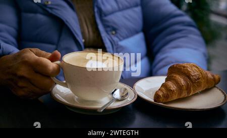 Frau, die eine Tasse Cappuccino und ein Croissant auf einem Teller auf dem Tisch hält Stockfoto