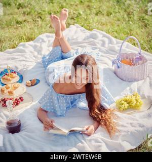 Picknickszene: Süßes lächelndes Mädchen, das auf einem Karo liegt und im Garten Buch liest. Außenporträt einer schönen glücklichen jungen Frau in blauem Kleid Stockfoto