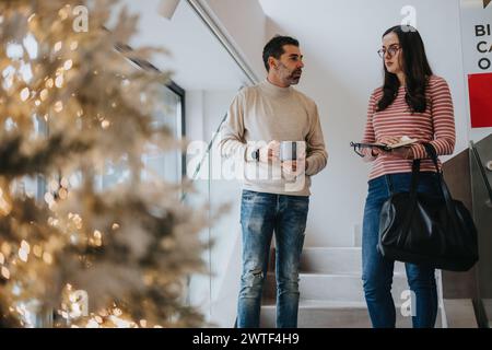 Zwei Senioren, die sich an einer zwanglosen Diskussion an einem geschmückten Weihnachtsbaum in einer modernen Büroumgebung beteiligen. Stockfoto