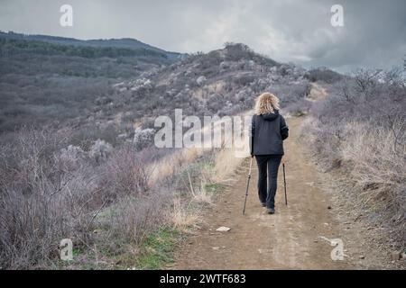 Blonde Erwachsene Frau, die Nordic Walking auf einem Pfad in den Bergen macht, bewölktes Frühlingswetter, freier Platz für eine aktive Lifestyle-Idee Stockfoto