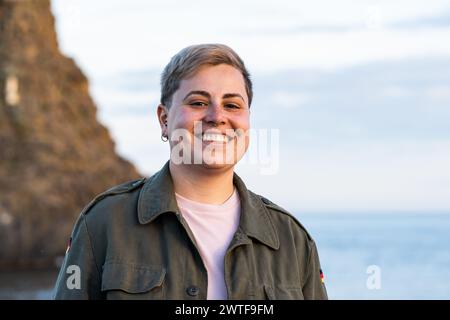 Junge Erwachsene geschlechtsneutral mit kurzen Haaren - fröhliches Lächeln vor dem Hintergrund des Meeres - ruhige Outdoor-Momente in der Abenddämmerung. Stockfoto