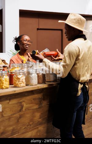 Ein umweltfreundlicher afroamerikanischer männlicher Ladenbesitzer verkauft frische Lebensmittel aus biologischem Anbau. Junge weibliche Kundin fragt nach nahrhaften Produkten aus der Region im kunststofffreien Laden. Stockfoto