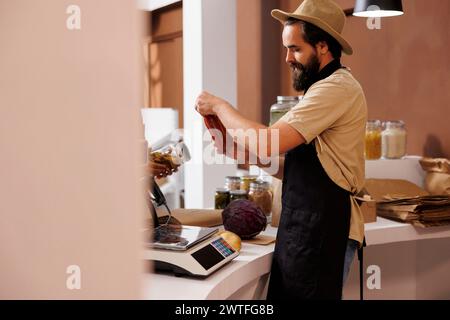 Der männliche kaukasische Ladenbesitzer, der einen stilvollen Hut und eine schwarze Schürze trägt, steht hinter der Kasse und hält ein Glas mit Nudelsauce. Anbieter analysiert Glasbehälter am Kassenschalter. Stockfoto