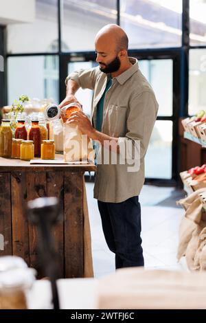 Ein Mann aus dem Nahen Osten füllt einen braunen Papierbeutel mit orangen Linsen. Junger Mann, der Körner in Beutel gießt, gesunde Waren aus wiederverwendbaren Behältern mit organischen Schüttgütern kauft. Stockfoto