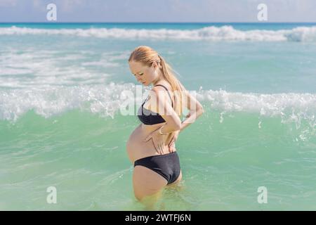 Im Meer erlebt eine schwangere Frau einen Moment des Unbehagens, während sie steht, sanft um ihren Rücken kümmert und die Herausforderungen von Rückenschmerzen unterstreicht Stockfoto