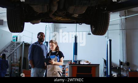 Qualifizierter Techniker in der Werkstatt zeigt dem Kunden, was an seinem Fahrzeug geändert werden muss. Garagenangestellter inspiziert das Fahrzeug auf einem Deckenlift und spricht mit einem afroamerikanischen Kunden Stockfoto