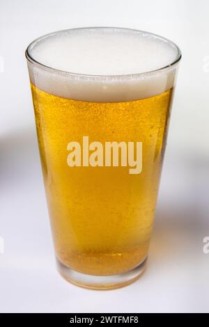 Ein Glas Bier sitzt auf weißem Hintergrund. Das Glas ist halb voll und das Bier ist schaumig Stockfoto