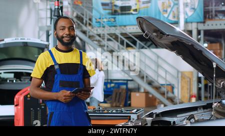 Porträt eines lächelnden Mechanikers im Autoservice, der mit einem Tablet neue Teile für beschädigte Fahrzeuge bestellt. afroamerikanischer Reparaturmann, der online nach Komponenten sucht, die alte in einem kaputten Auto ersetzen können Stockfoto