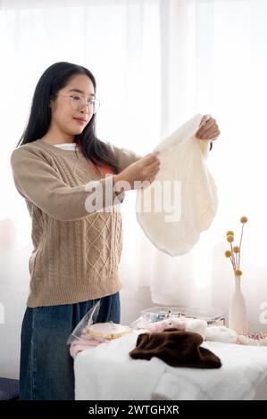 Eine positive asiatische Frau, die Stoff auf ihre Handwerkskleidung überprüft und Nähzubehör am Tisch vorbereitet. Handarbeit, Stickerei, Schneider Stockfoto