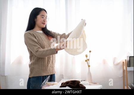 Eine positive asiatische Frau, die Stoff auf ihre Handwerkskleidung überprüft und Nähzubehör am Tisch vorbereitet. Handarbeit, Stickerei, Schneider Stockfoto