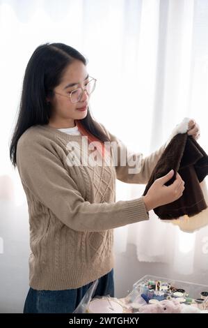 Eine positive asiatische Frau, die Stoff auf ihre Handwerkskleidung überprüft und Nähzubehör am Tisch vorbereitet. Handarbeit, Stickerei, Schneider Stockfoto