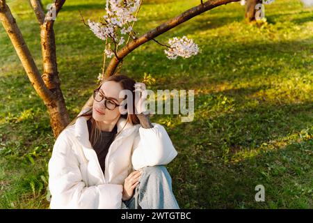 Eine schöne junge Frau in Brille und weißem Mantel sitzt neben einem blühenden Baum Stockfoto