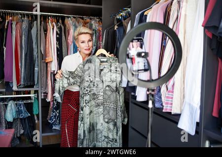 Schöne reife Frau, Stylistin Social Media Influencer und Video Bloggerin, nimmt auf ihrem Handy ein Video über Kleidung und Mode in einem Kleiderschrank auf Stockfoto
