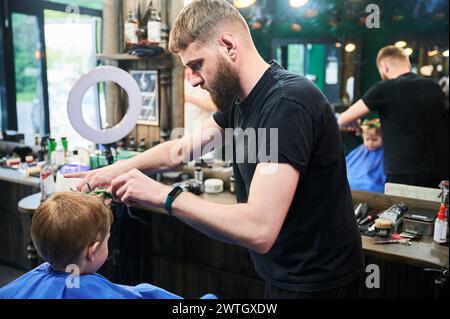 Ein professioneller Friseur macht Friseur für seinen kleinen Kunden. Männlicher Friseur mit Friseurwerkzeugen. Rückansicht eines kleinen Jungen, der seinen ersten Haarschnitt bekam. Stockfoto