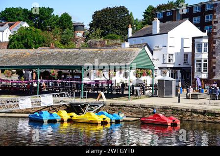 Pedalos, die am Fluss exe verankert sind, mit Geschäften und Restaurants entlang des East Quay im Hinterland, Exeter, Devon, Großbritannien, Europa. Stockfoto