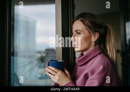 Frau genießt eine Tasse heißen Tee, Kaffee zu Hause nach der Arbeit, steht am Fenster und trägt einen rosa Hoodie. Work-Life-Balance für das Pflegepersonal. Stockfoto