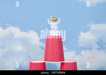Eine weiße Astronautenfigur mit goldenem Helm auf einer Pyramide aus roten Pappbechern, vor einem Hintergrund des blauen Sommerhimmels mit weißen Wolken. Leuchtende Farben Stockfoto