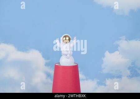 Eine weiße Astronautenfigur mit goldenem Helm auf einer Pyramide aus roten Pappbechern, vor einem Hintergrund des blauen Sommerhimmels mit weißen Wolken. Leuchtende Farben Stockfoto