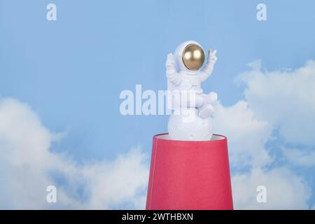 Eine weiße Astronautenfigur mit goldenem Helm auf einer Pyramide aus roten Pappbechern, vor einem Hintergrund des blauen Sommerhimmels mit weißen Wolken. Leuchtende Farben Stockfoto