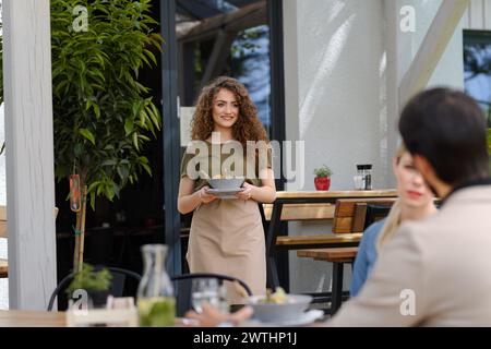 Porträt einer schönen Kellnerin, die einen Teller mit Essen hält, eine Schüssel mit Salat. Server im Vorfeld bringt Speisen zu den Gästen auf der Restaurantterrasse. Stockfoto